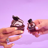 Two hands holding Vegan Red Velvet Volcano Cupcakes with Oreo and ganache topping on purple background